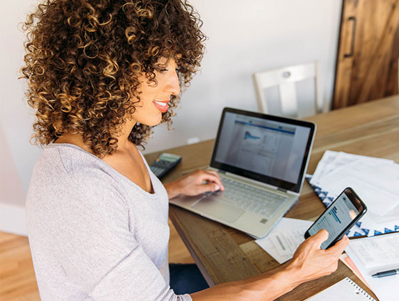 young woman sitting at kitchen table looking at cell phone and laptop of online banking report