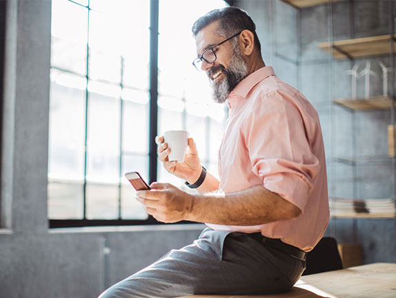 man sitting on table in industrial style office holding cell phone and coffee cup